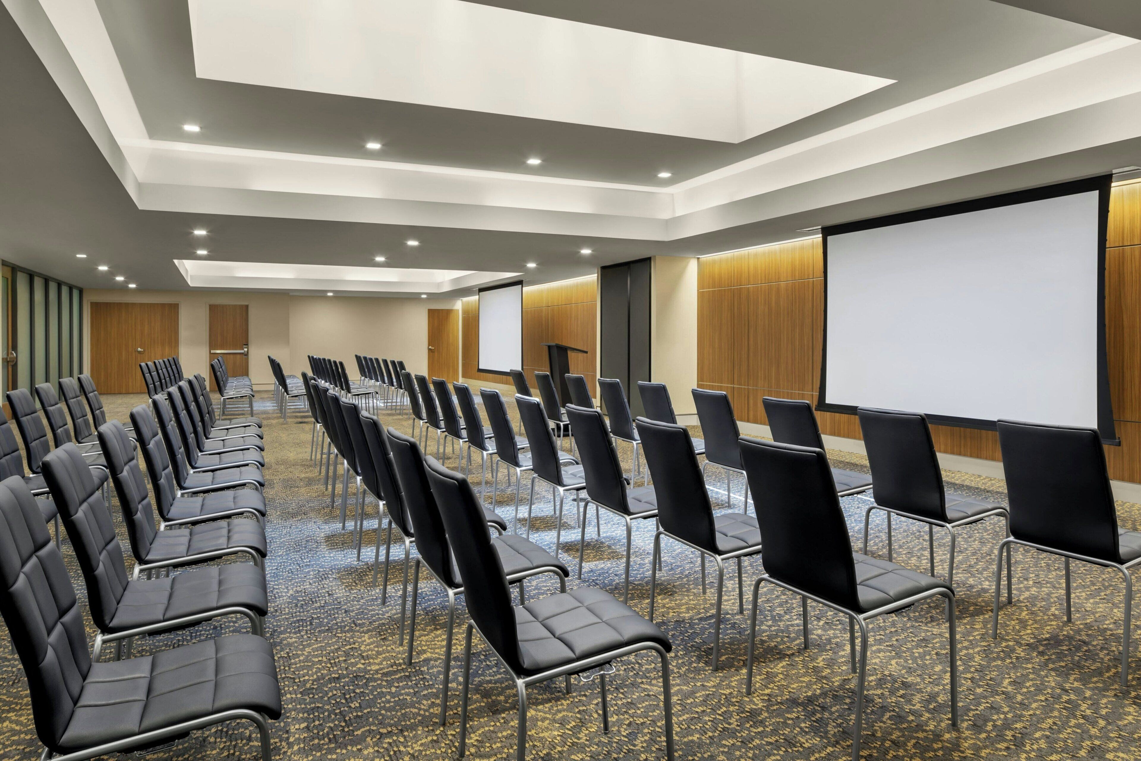 A conference room with rows of black chairs, a large screen, and a podium.