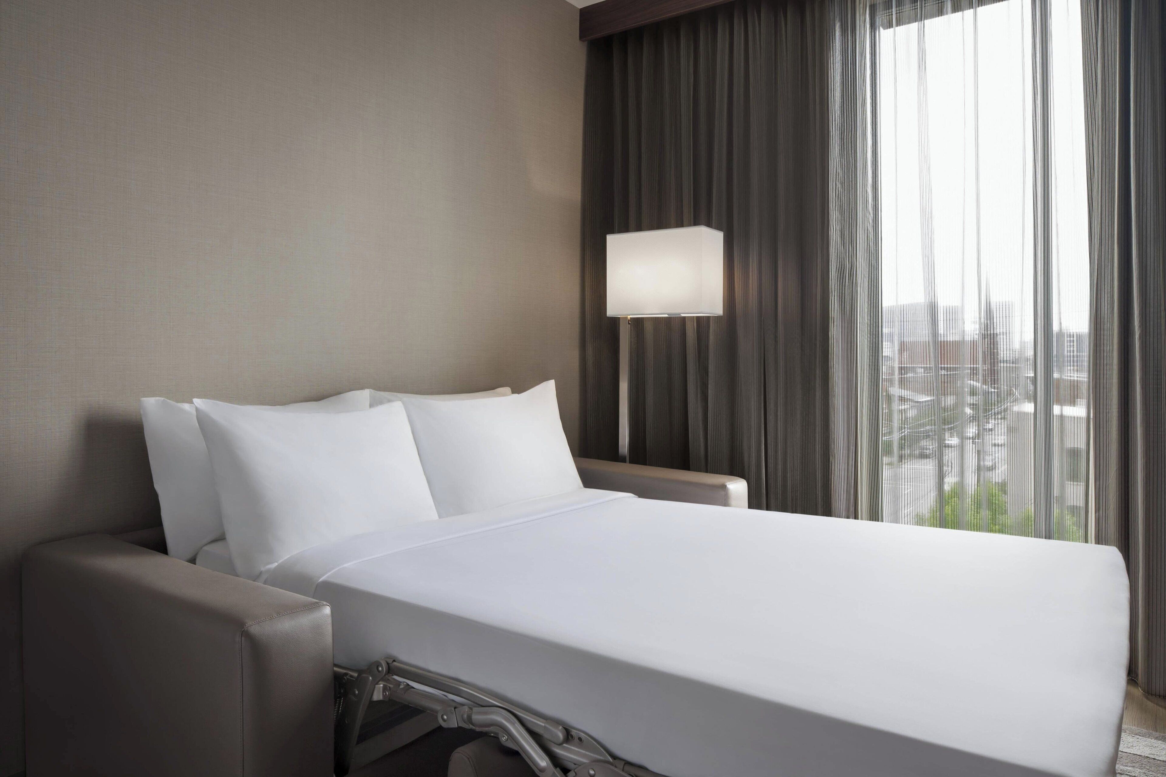 A hotel room with a large bed, a bedside table, a lamp, and a view of the city through the window.
