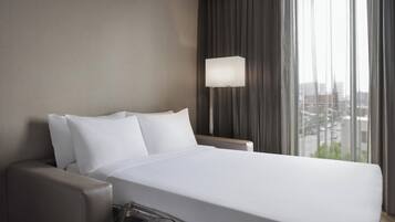A hotel room with a large bed, a bedside table, a lamp, and a view of the city through the window.