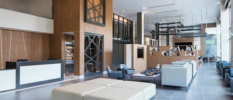 A modern hotel lobby with a reception desk, seating area, and a large window providing natural light.