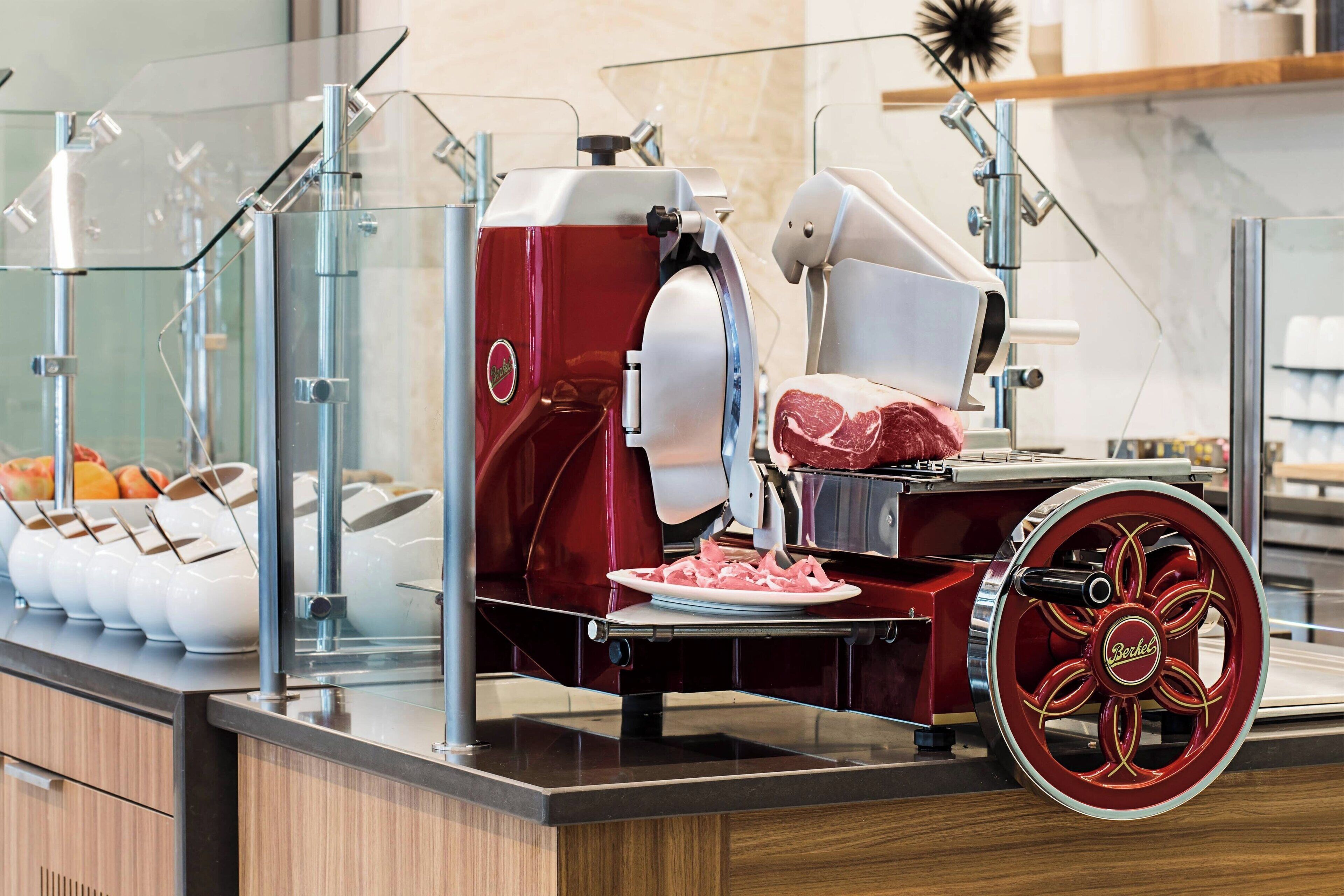 A meat carving station with a red and white machine, a plate of sliced meat, and a glass display case with bowls.
