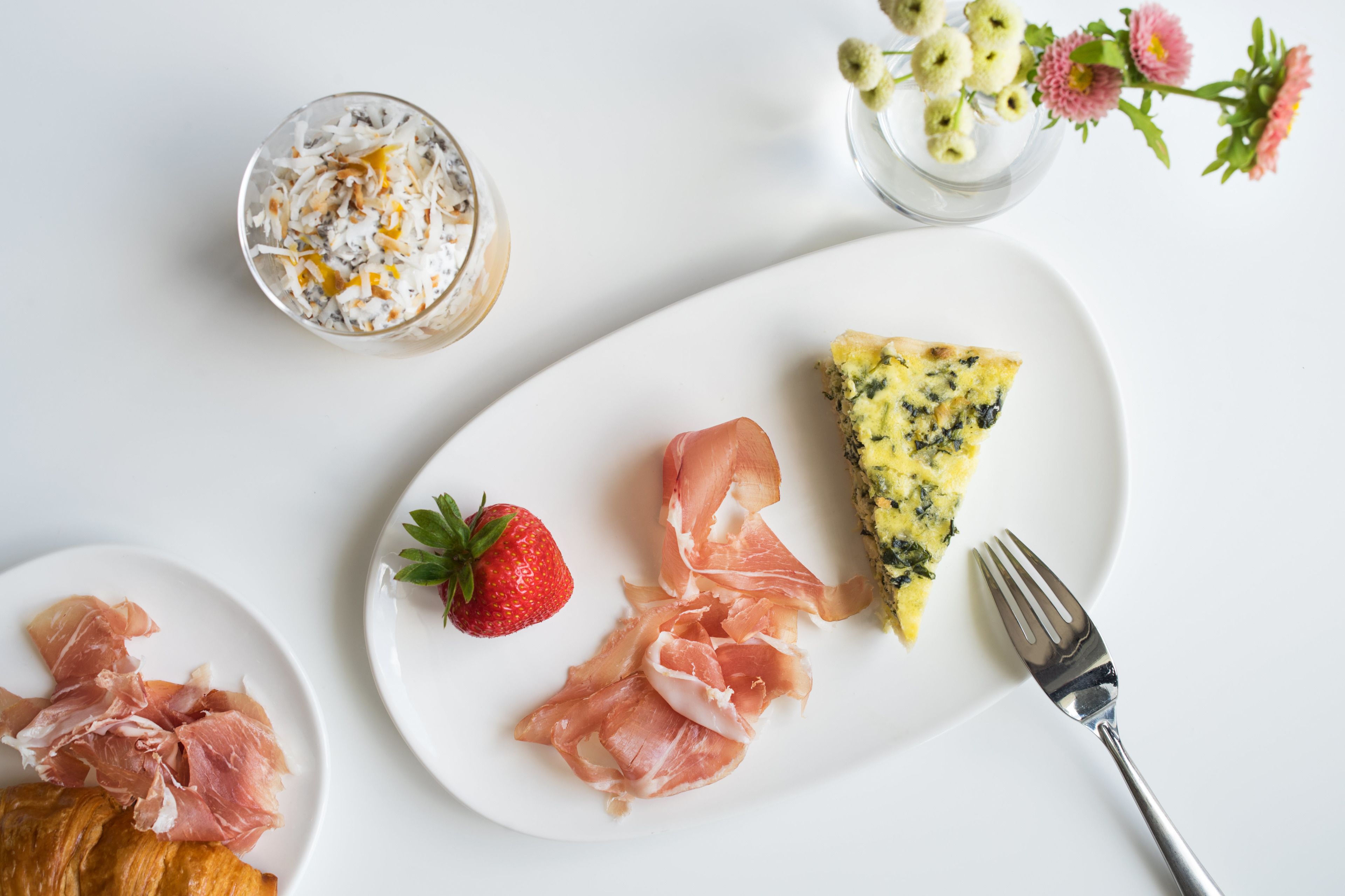Een ontbijttafel met een bord eten, inclusief een plakje quiche, parmaham en een aardbei, plus een glas romige drank en een croissant.