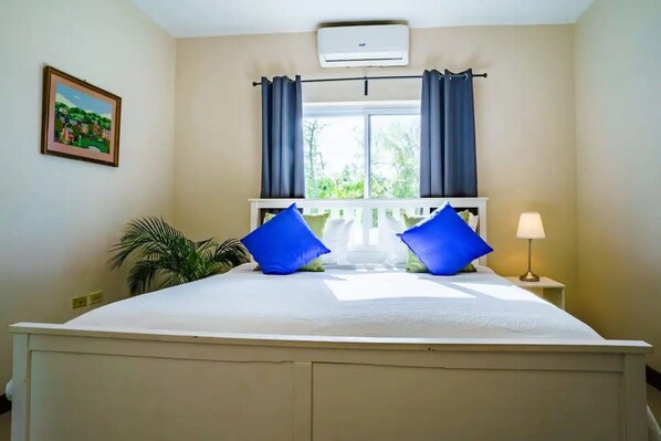 A bedroom with a large bed, two blue pillows, a nightstand with a lamp, a window with curtains, an air conditioner, and a framed picture on the wall.