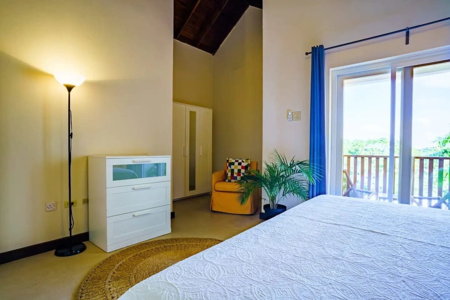 A bedroom with a bed, a dresser, a chair, a potted plant, and a balcony with a railing.