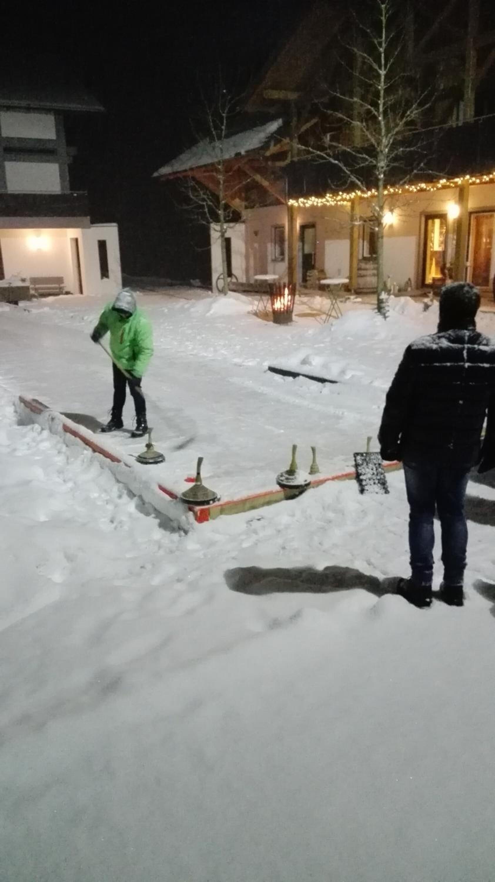 A snowy outdoor area with a fire pit and tables, a building with lights, and a person in a green jacket.
