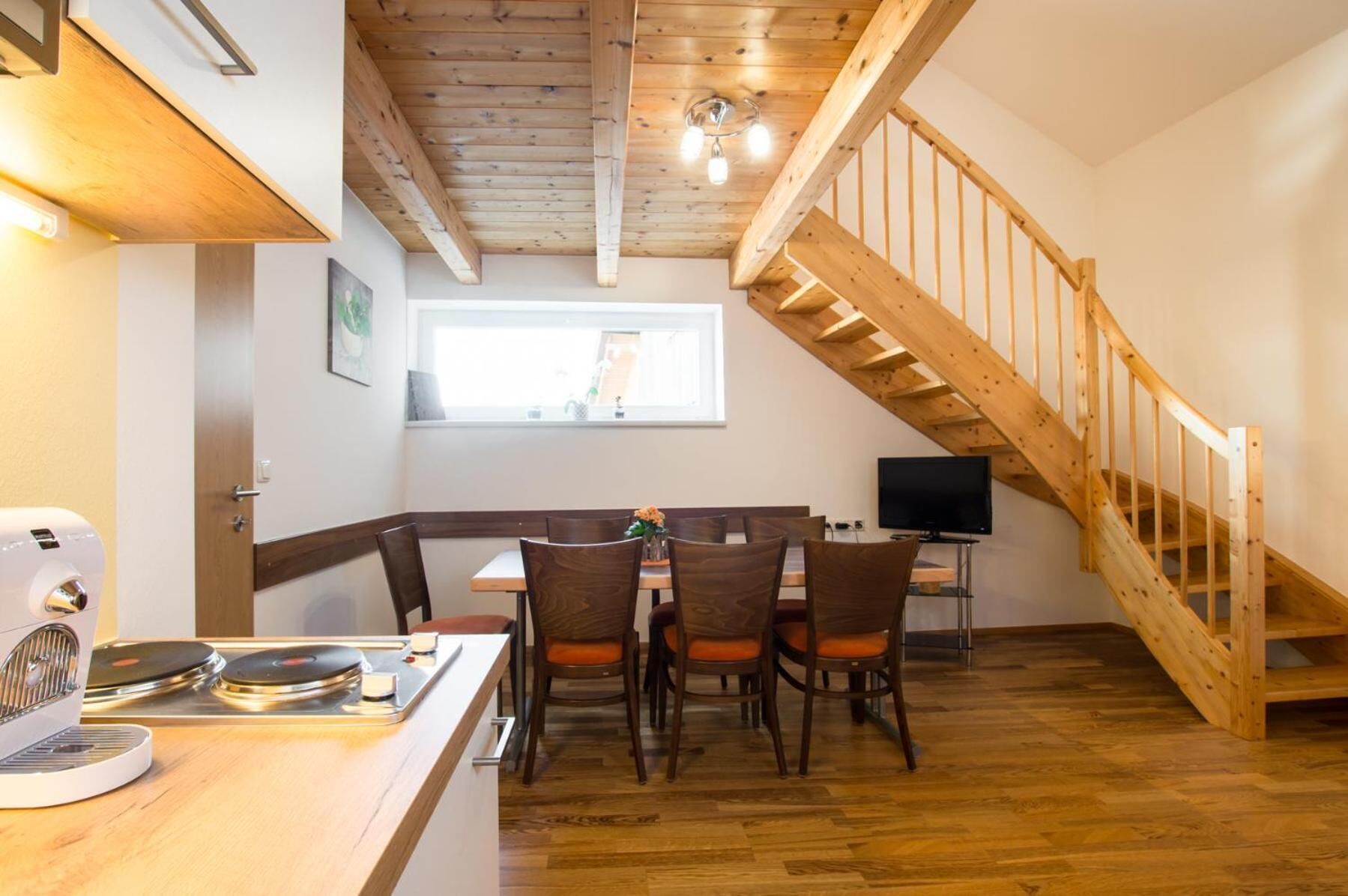 A cozy kitchen with wooden flooring, a dining area with chairs, a coffee machine, and a TV on a stand.