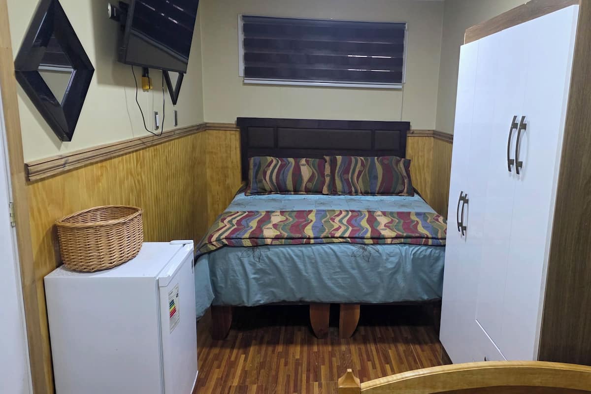 A small room with a bed, a television mounted on the wall, a window with blinds, a wooden floor, and a wardrobe.