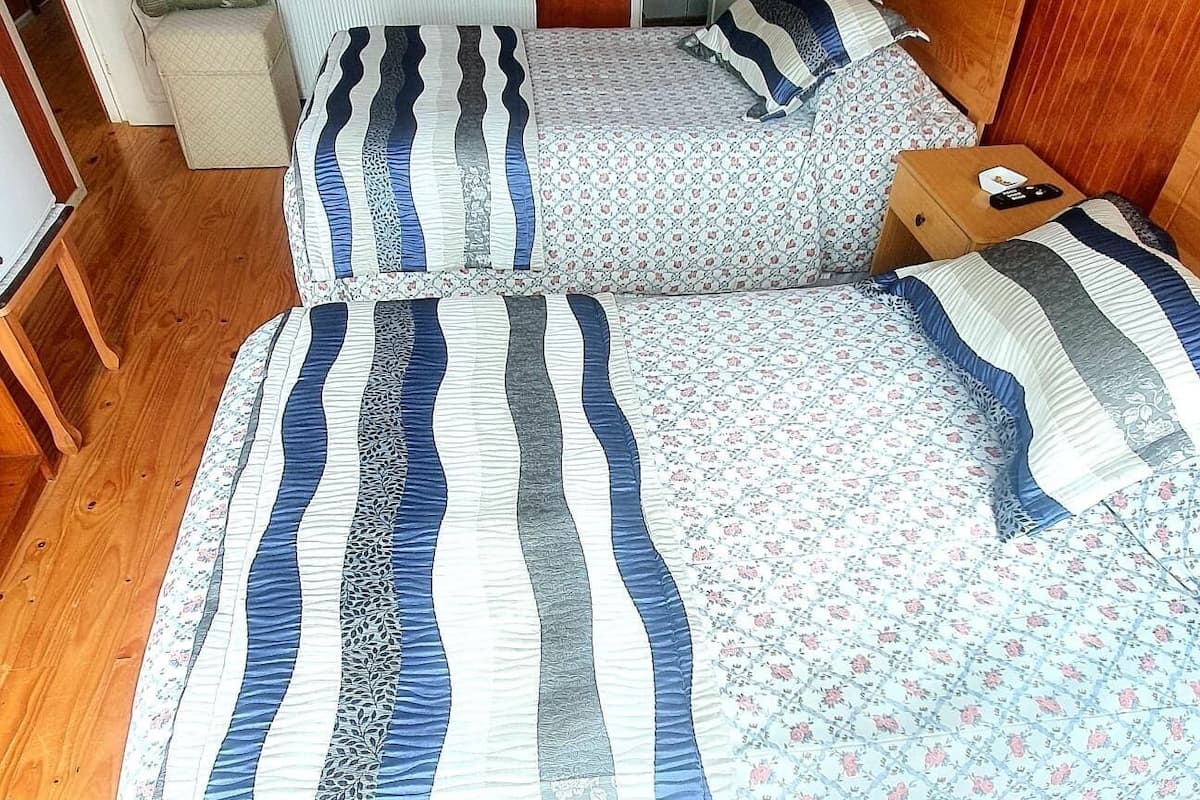 A room with two beds, a wooden floor, a radiator, a wooden door, and a small wooden nightstand with a remote control.