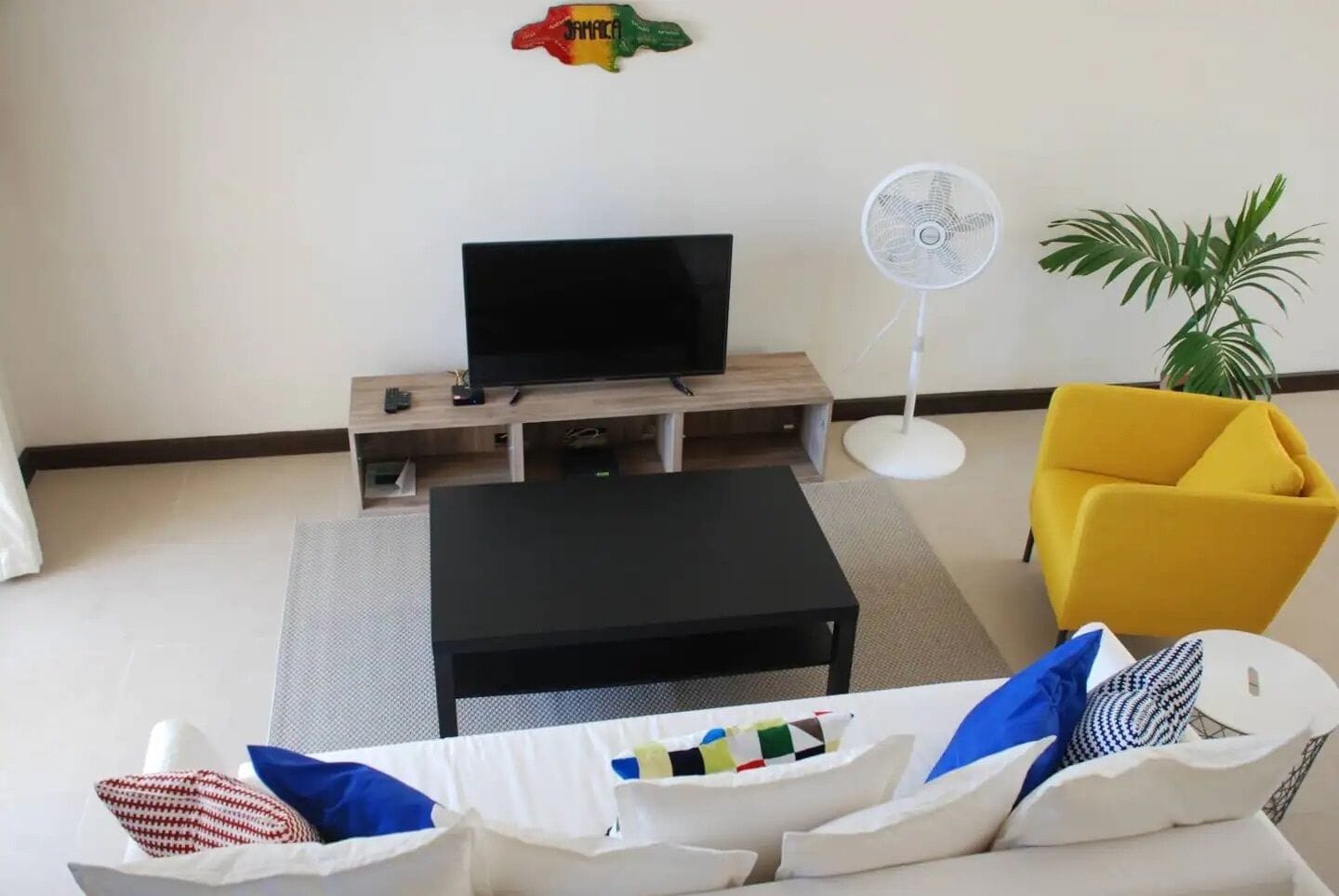 Una sala de estar con un televisor sobre un mueble de madera, un sofá blanco con almohadones de colores, un sillón amarillo, un ventilador de pie blanco y una planta en maceta.