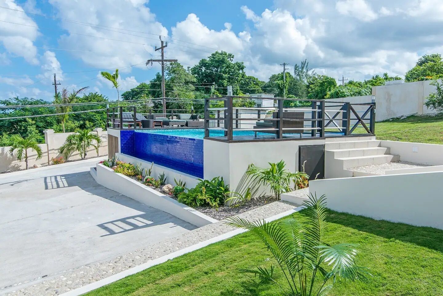A modern outdoor pool area with a blue-tiled section, surrounded by a white wall, and a staircase leading up to a deck with lounge chairs.
