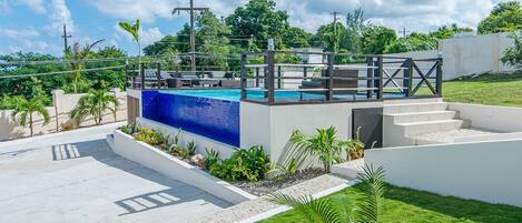 A modern outdoor swimming pool with a blue water feature, surrounded by a white deck and steps leading up to a covered seating area.