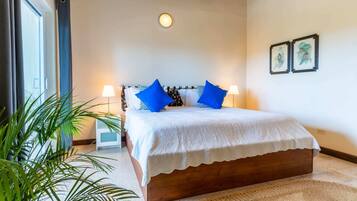 A bedroom with a wooden bed, white bedding, two blue pillows, a bedside lamp, a large window with curtains, and a wall-mounted picture.