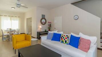 Sala de estar com sofá branco, poltrona amarela e mesa de centro preta. É possível ver, ao fundo, uma área de jantar com uma mesa e cadeiras.
