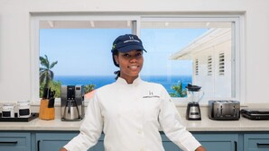 A person in a white chef's coat with "Hospitality Expert" on the chest, standing in a kitchen with a view of the ocean through a large window. - Hospitality Expert CB4 (Montego Bay)