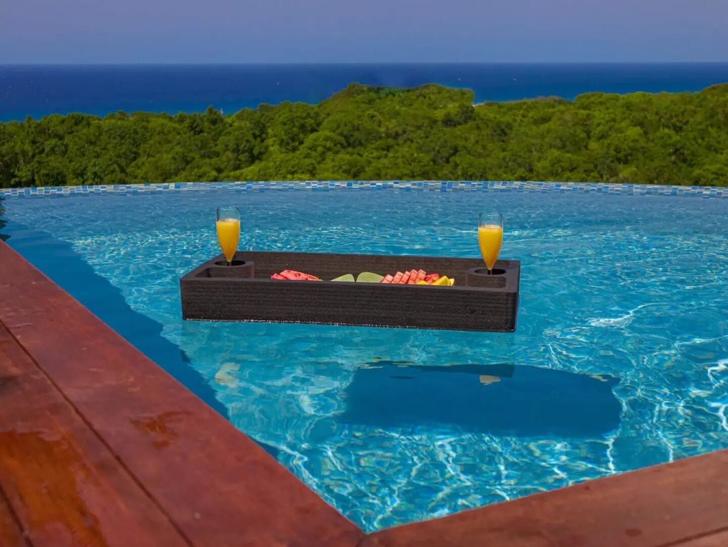 A pool with a floating bar and drinks, surrounded by a wooden deck and a view of the ocean.