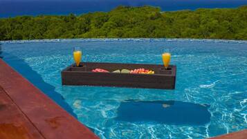 A pool with a floating bar and drinks, surrounded by a wooden deck and a view of the ocean.