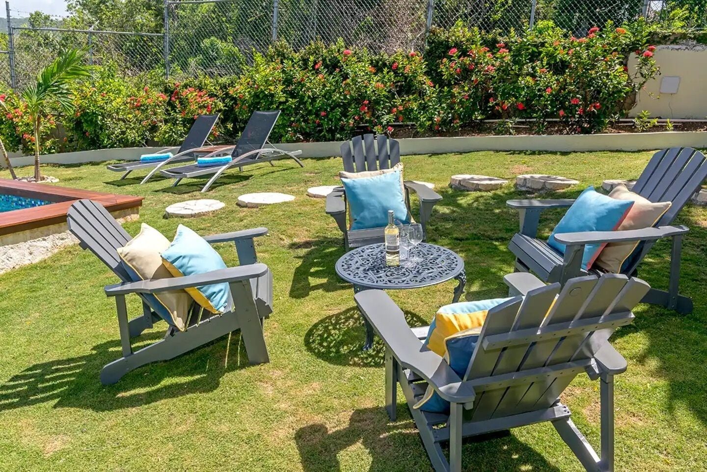 An outdoor seating area with Adirondack chairs and a round table, surrounded by a well-maintained lawn and flowering bushes.