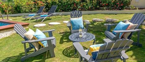 An outdoor seating area with Adirondack chairs and a round table, surrounded by a well-maintained lawn and flowering bushes.