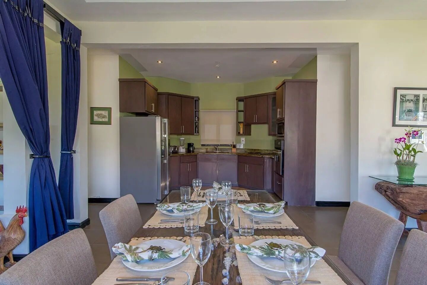 Comedor con una mesa dispuesta para seis, cocina al fondo y una cortina azul a la izquierda.