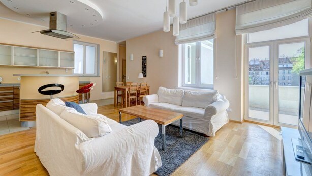 A modern living room with a sofa, a coffee table, and a TV. There is a dining area with chairs and a table. The room has large windows and a view of buildings outside. - Apartament Opera Sopot (Sopot)