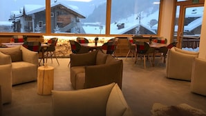 A cozy lounge area with sofas and chairs arranged around a central table, large windows offering a snowy mountain view. - Hotel Cristiania (Pinzolo)