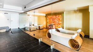 A modern spa area with a glass wall, wooden flooring, and a white relaxation area with a massage table. - Hotel Cristiania (Pinzolo)