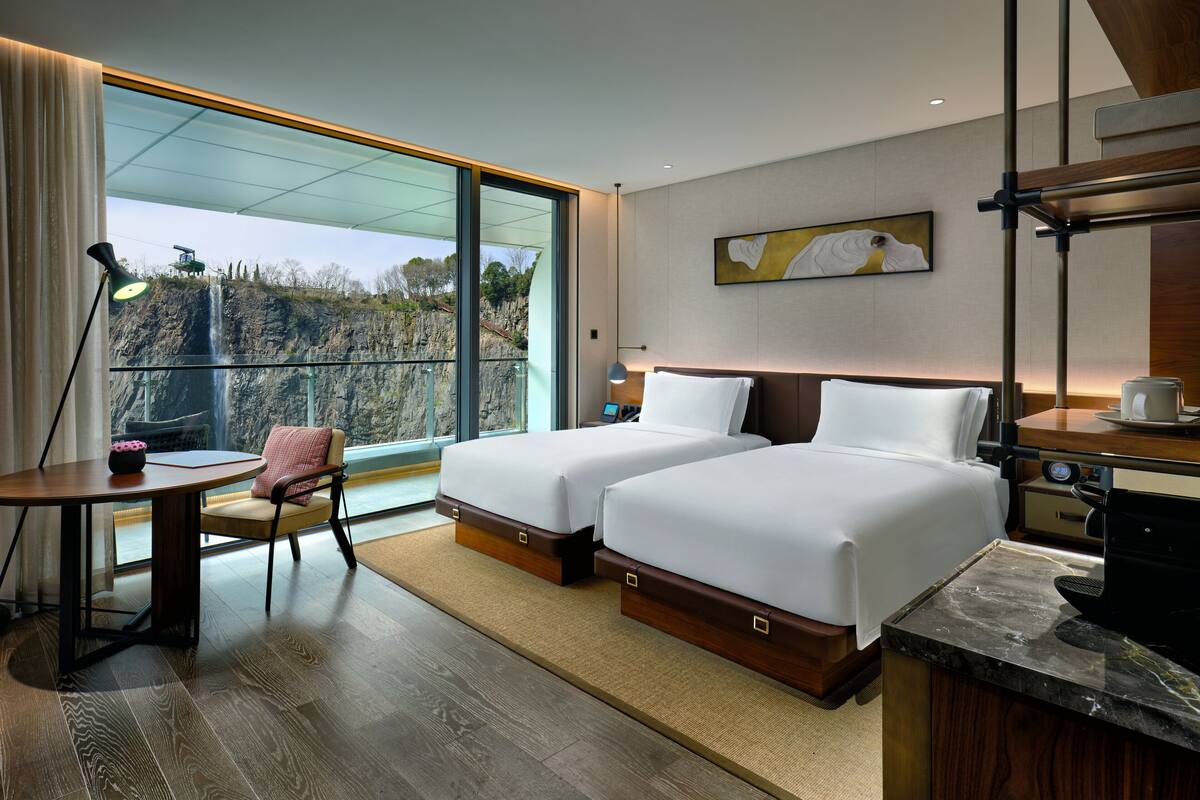 A hotel room with two beds, a desk, a chair, a large window with a view of a cliff, and a painting on the wall.