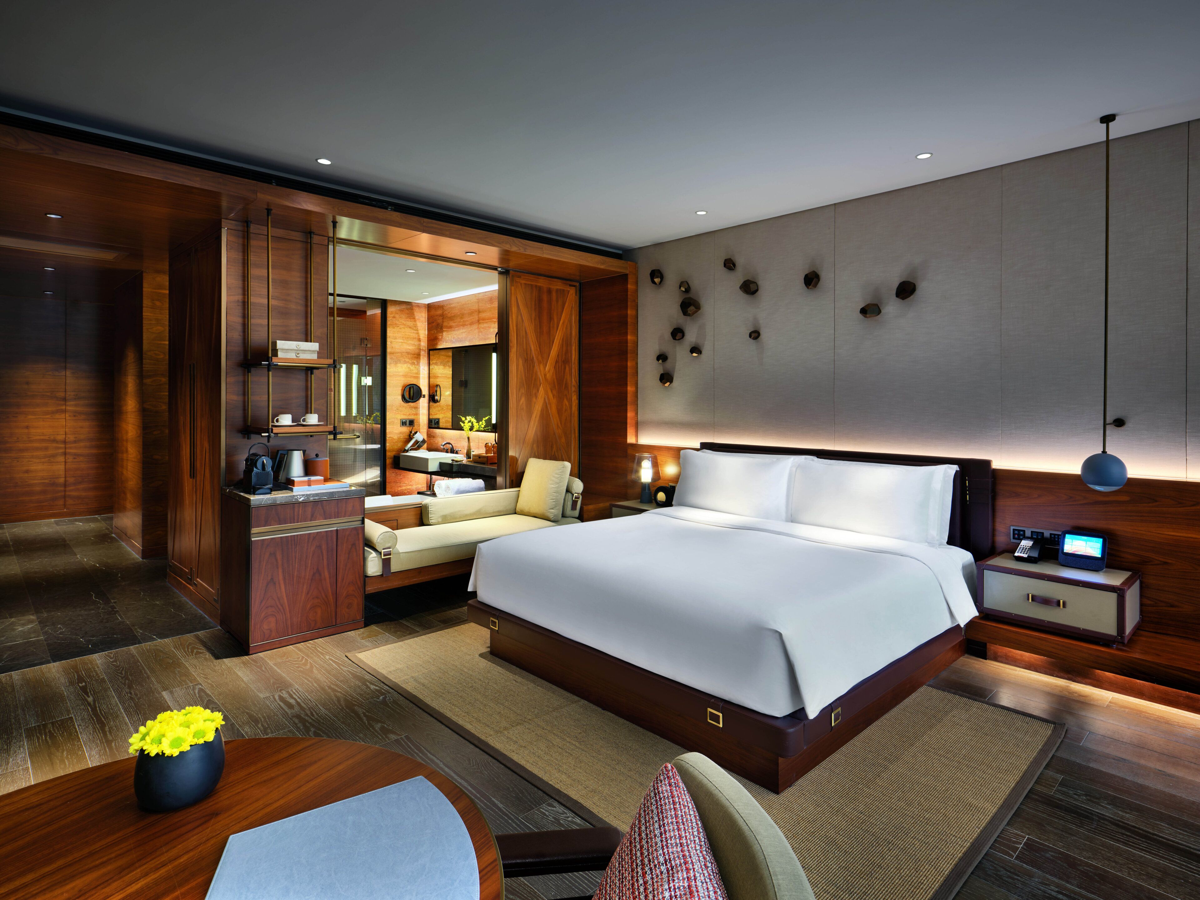 A modern hotel room with a large bed, a desk, a sofa, a vanity with a mirror, and a bathroom visible through an open door.