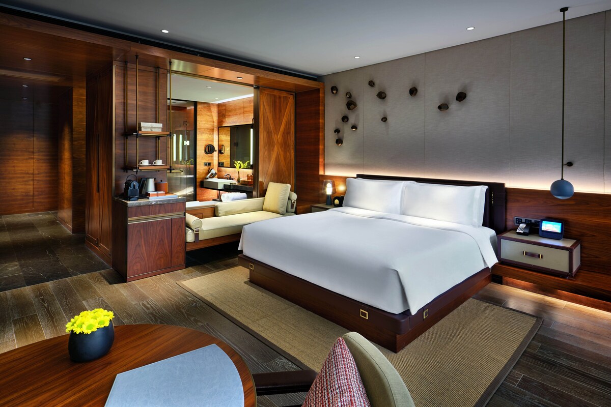 A modern hotel room with a large bed, a desk, a sofa, a vanity with a mirror, and a bathroom visible through an open door.