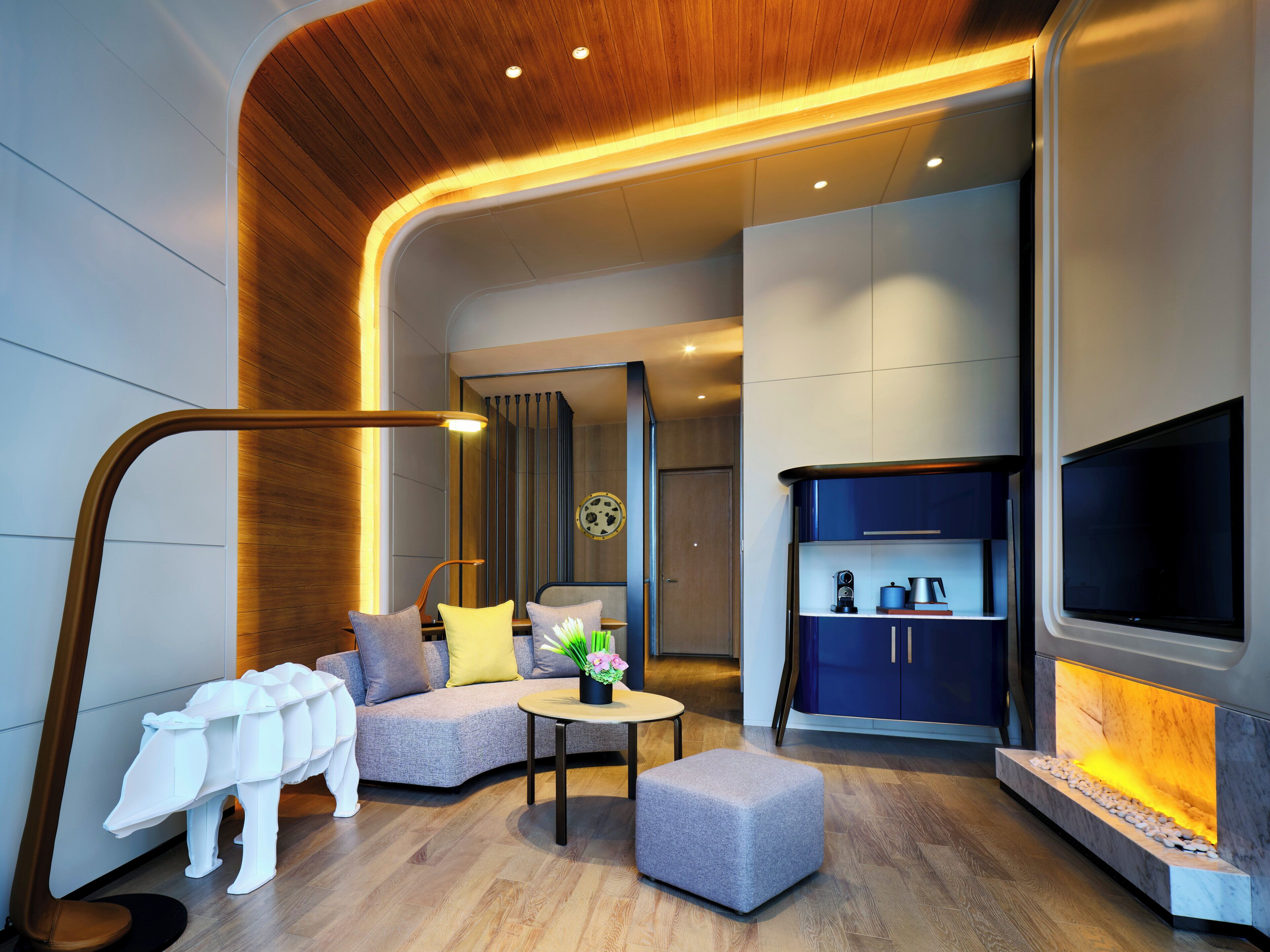 A modern living room with a sofa, ottoman, and a coffee table. There is a TV mounted on the wall and a fireplace with a stone surround.