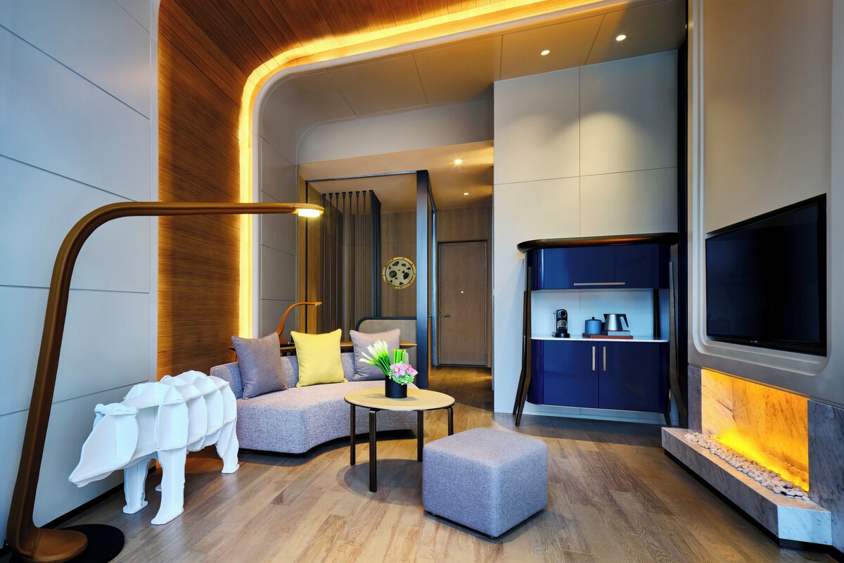 A modern living room with a sofa, ottoman, and a coffee table. There is a TV mounted on the wall and a fireplace with a stone surround.