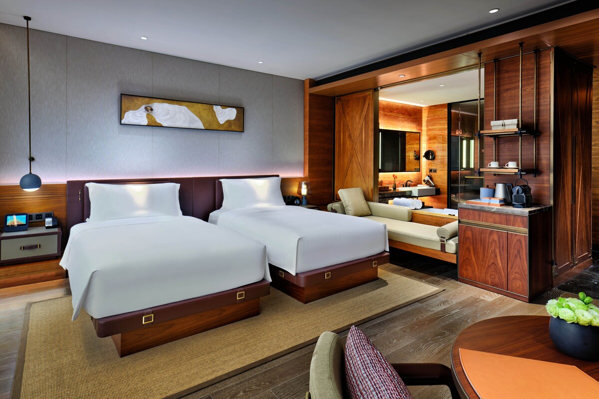 A modern hotel room with two beds, a sitting area, a dining table, and a bathroom with a vanity and mirror.