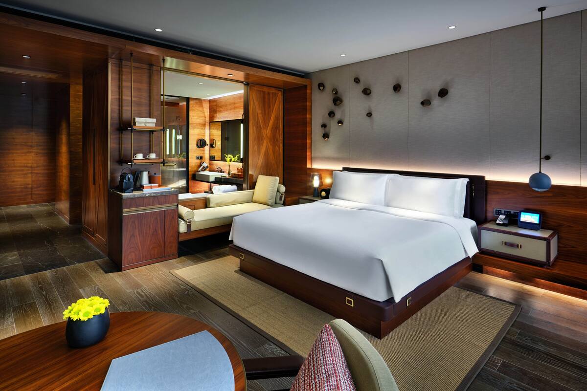 A modern hotel room with a large bed, a desk, a sofa, a vanity with a mirror, and a bathroom visible through an open door.