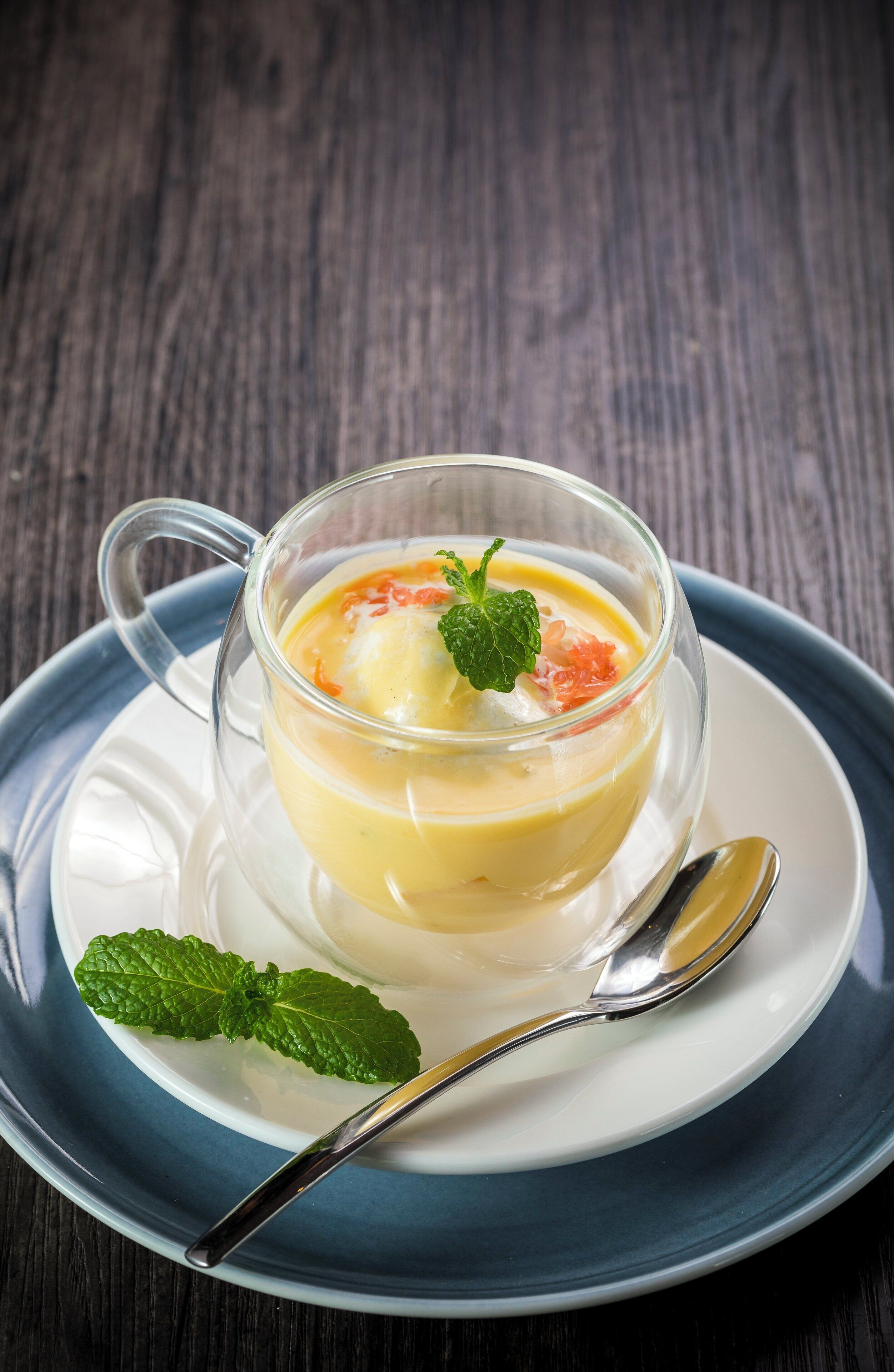A clear glass cup of soup with a spoon on a white plate, placed on a blue saucer.