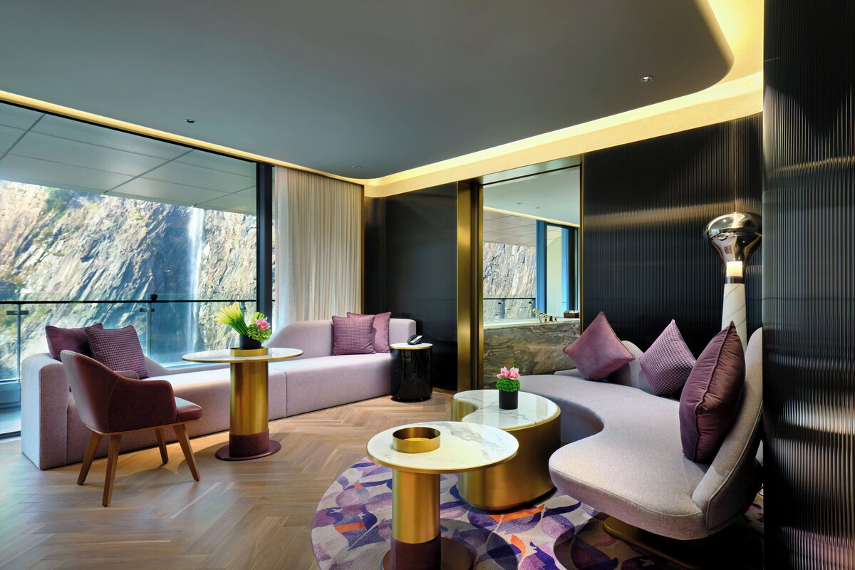 A modern hotel room with a large window offering a view of a waterfall, a sofa, a chair, and a coffee table.