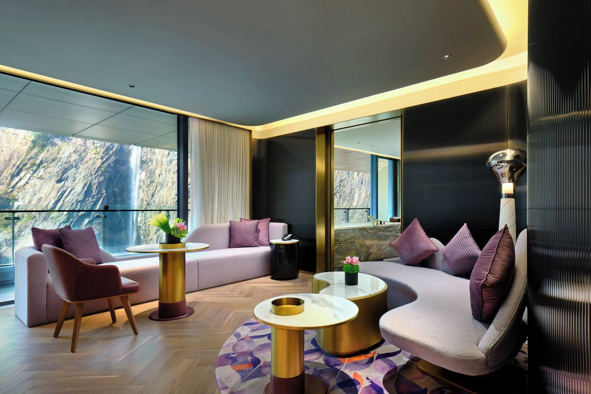 A modern hotel room with a large window offering a view of a waterfall, a sofa, a chair, and a coffee table.