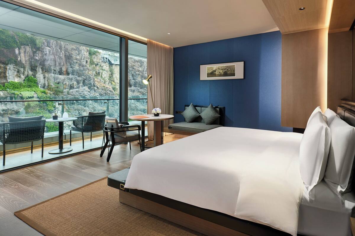 A modern hotel room with a large bed, a seating area with a table and chairs, a view of a rocky landscape through large windows, and a framed picture on the wall.