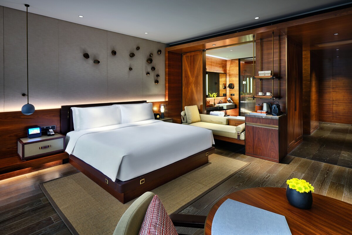 A modern hotel room with a large bed, a desk with a computer, a seating area, and a bathroom with a mirror and sink.
