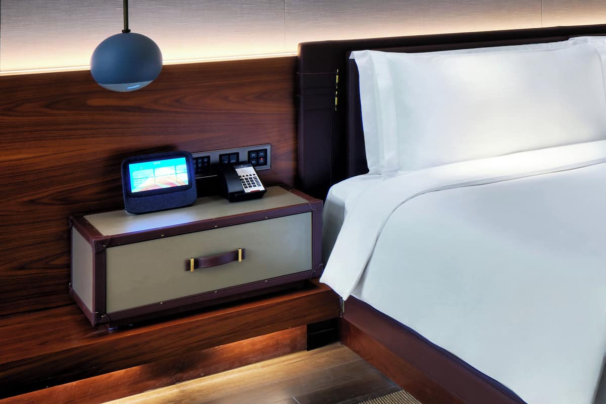 A neatly made bed with white linens, a bedside table with a lamp, a digital clock, and a phone.