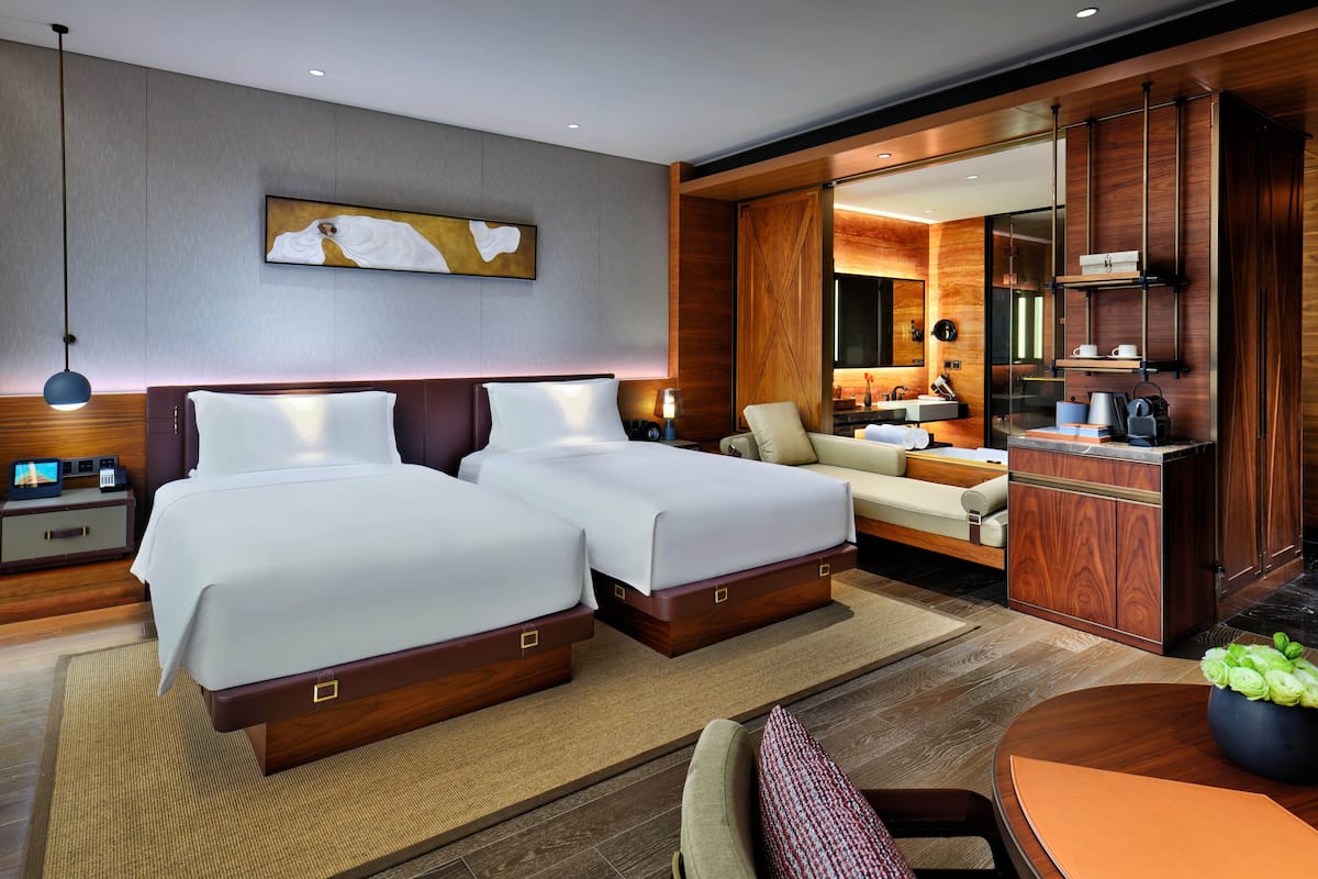 A modern hotel room with two beds, a sitting area, a dining table, and a bathroom with a vanity and mirror.