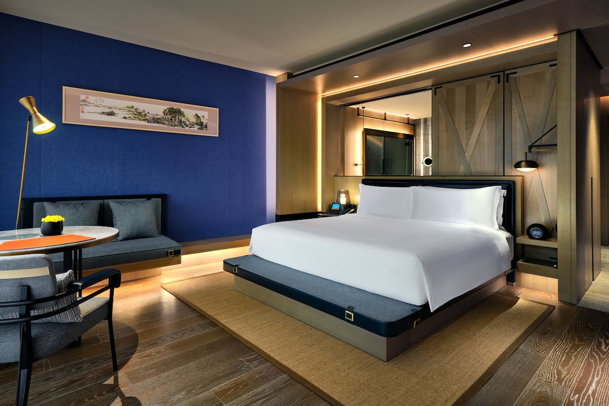 A modern hotel room with a large bed, a seating area with a table, and a wall-mounted TV.