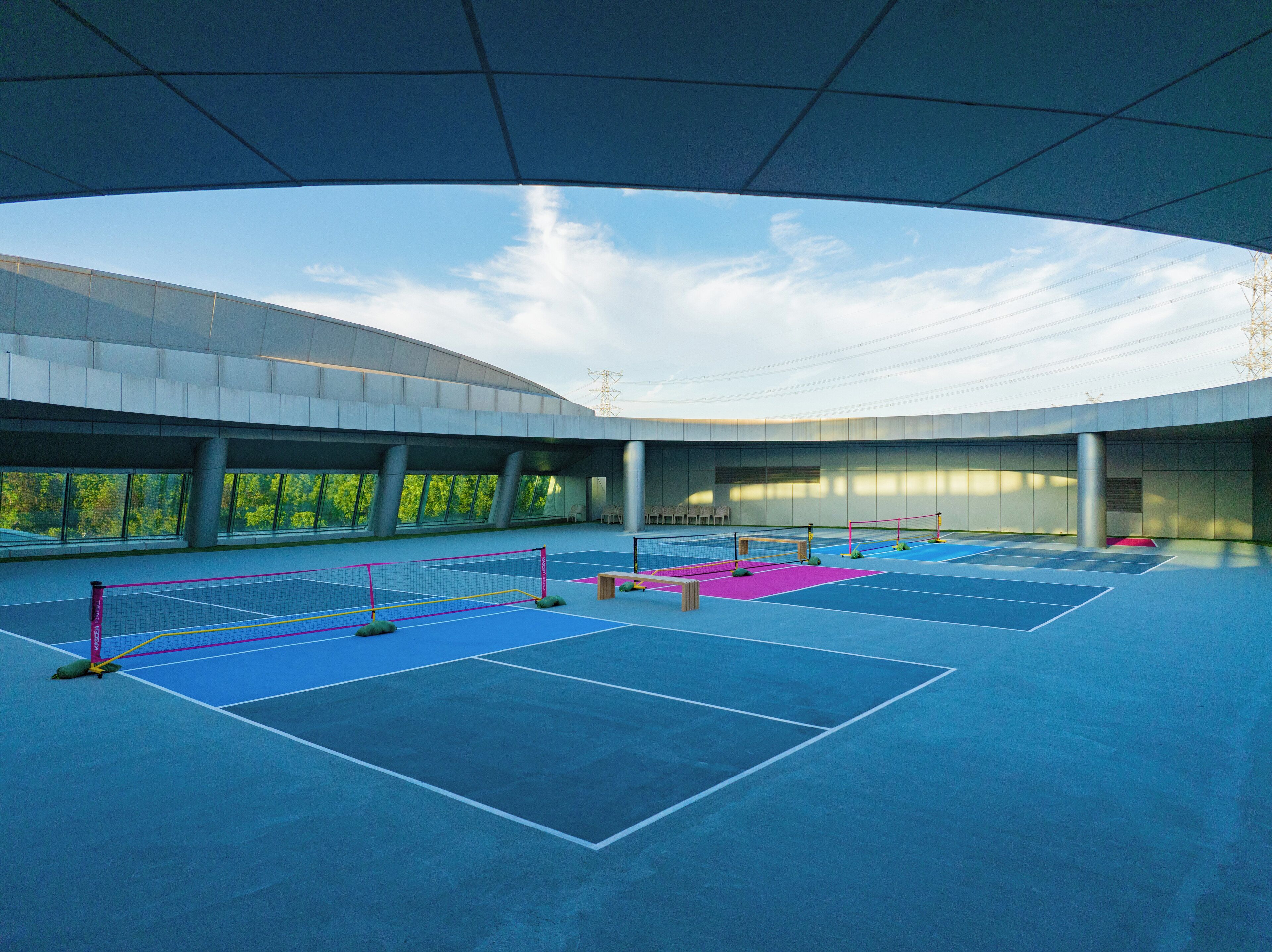 An indoor tennis court with multiple courts, seating areas, and a large glass wall providing a view of the outdoors.