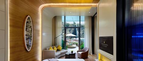 Une chambre d’hôtel moderne avec un plafond en bois, un grand lit, un coin salon avec un canapé et des fauteuils, une télévision et une vue sur une cascade à l’extérieur.