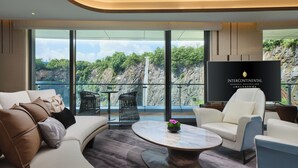 Een moderne hotelkamer met een groot raam dat uitzicht biedt op een waterval en groen. Er is een bank, een salontafel en een televisiescherm waarop de naam van het hotel staat.