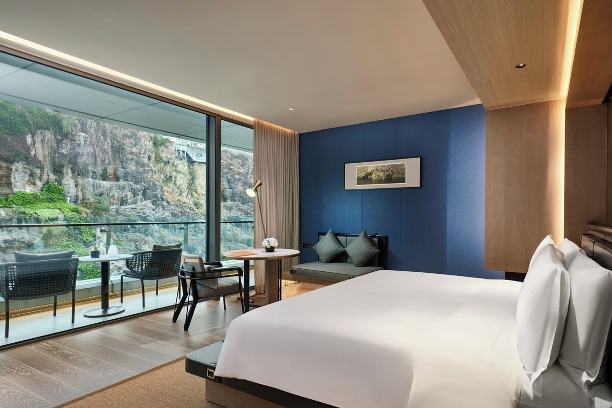 A modern hotel room with a large bed, a seating area with a table and chairs, a view of a rocky landscape through large windows, and a framed picture on the wall.
