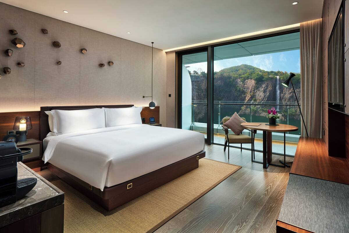 A modern hotel room with a large bed, a small table with a chair, a nightstand with a lamp, and a view of a waterfall through large windows.