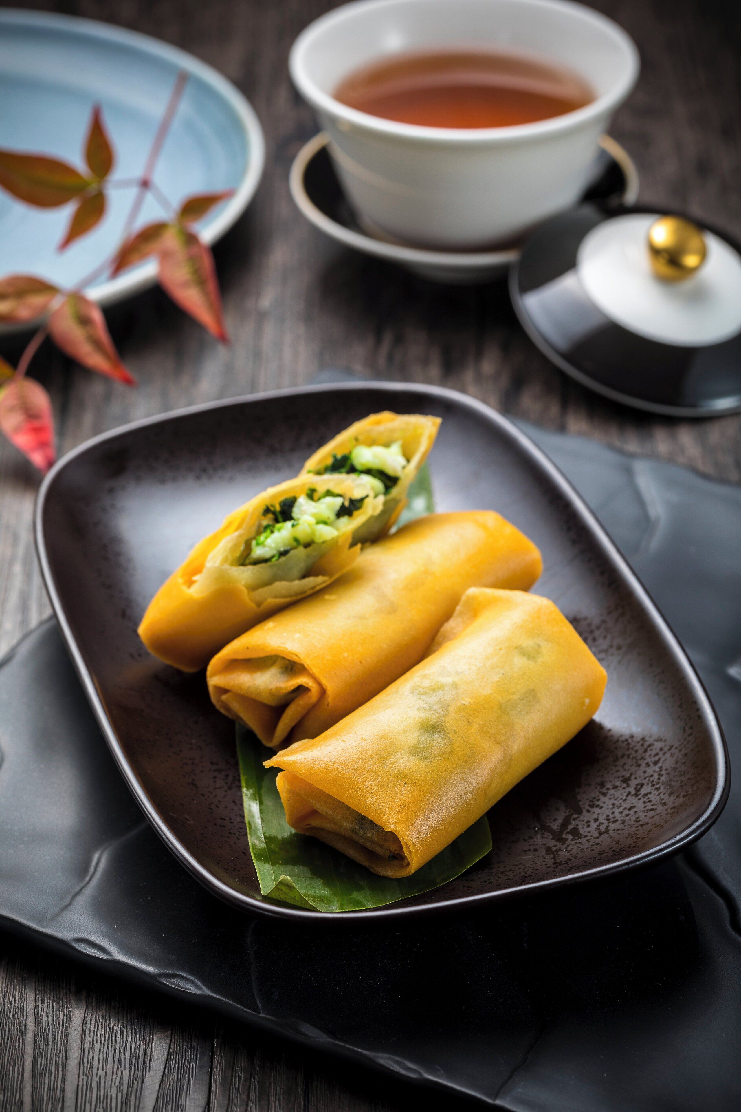Un plato de rollitos primavera sobre un plato oscuro, con una taza de té y un calentador de té al fondo.