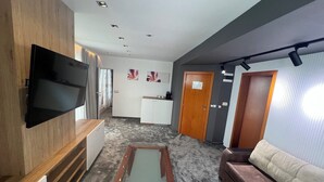 Un salon moderne avec un téléviseur à écran plat fixé à un mur en bois, un canapé marron, une table basse et un coin cuisine à l’arrière-plan.