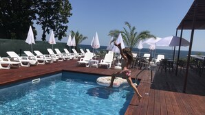Un espace piscine avec des chaises longues et des parasols, une terrasse en bois et une vue sur l’océan.