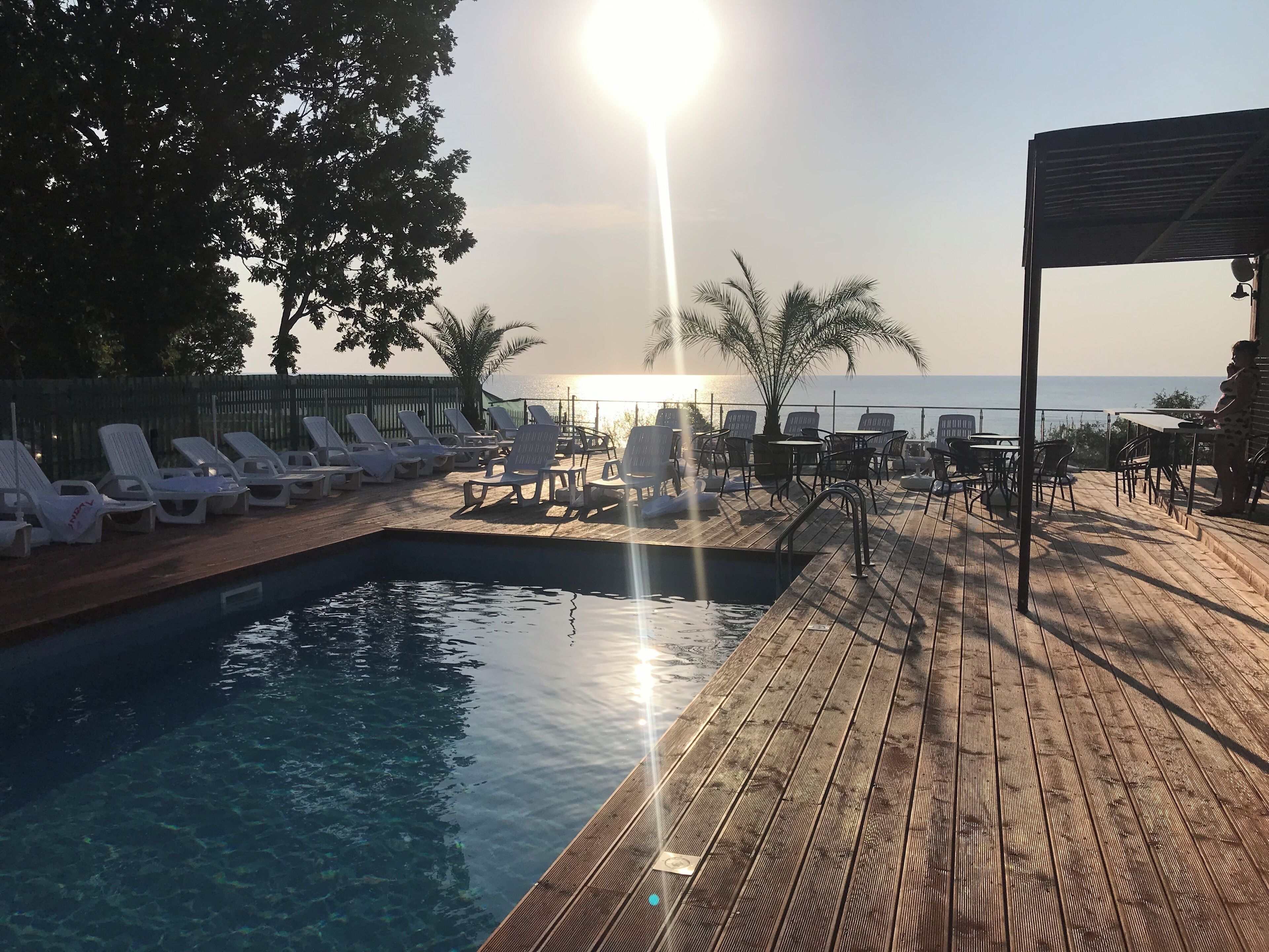 A wooden deck with a pool, lounge chairs, and tables overlooking a body of water.